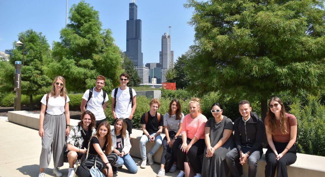 A group of students with backpacks sitting on a cement ledge with green trees and the Willis Tower in the background.
