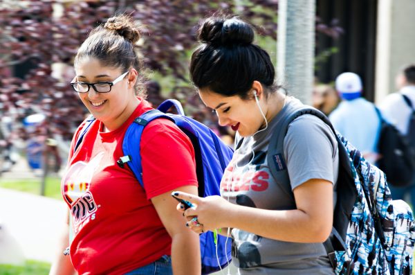 two students wearing UIC shirt with backpack and cellphone walking on campus