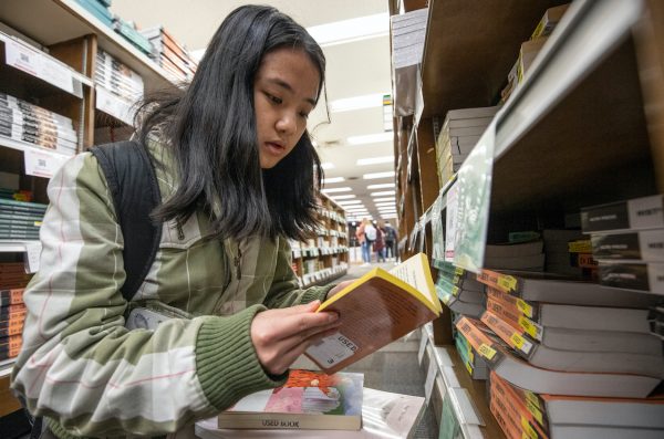 student looking at a used book with stack of books on bookstore shelf