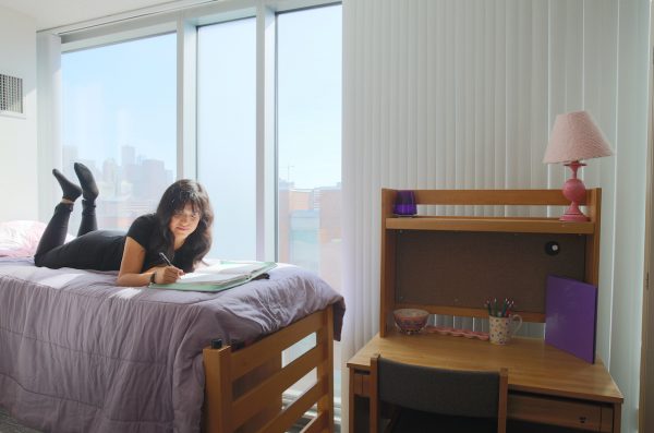 student reading a book lying on a bed  in a dorm bedroom with chair and work desk