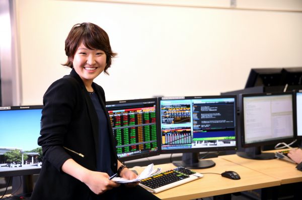 woman holding some papers standing in front of four computer monitors smiling at camera