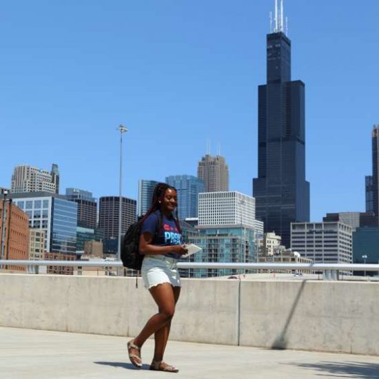 A UIC student wearing a backpack walks on the UIC campus with the Willis Tower in the background.