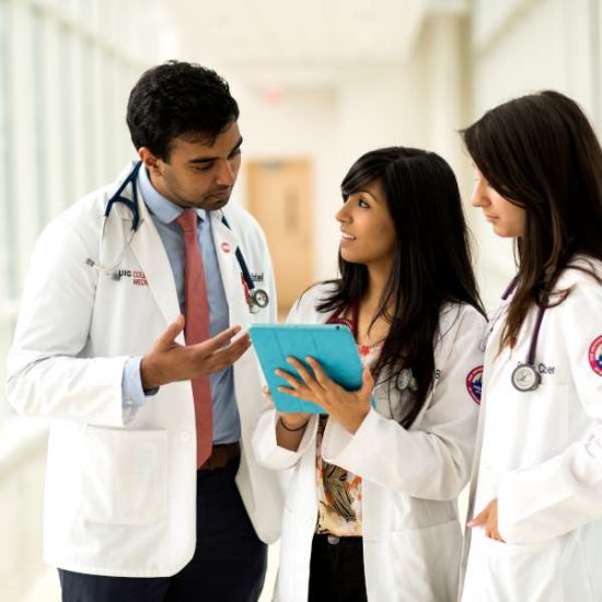 Three individuals in white medical coats discuss content on a tablet.