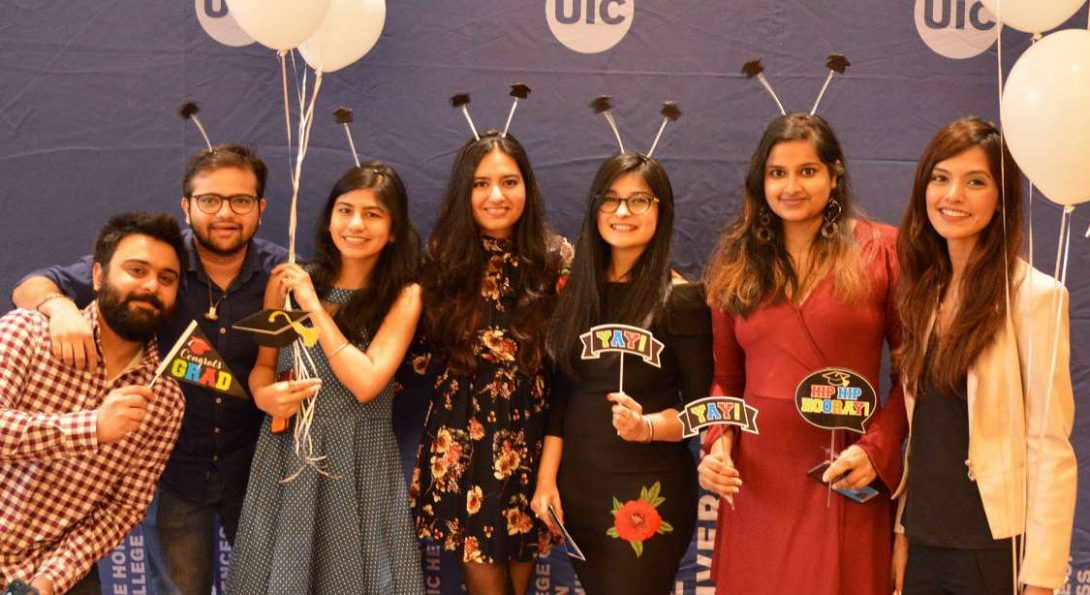 Seven graduating students stand holding white baloons, smiling in front of a UIC banner,