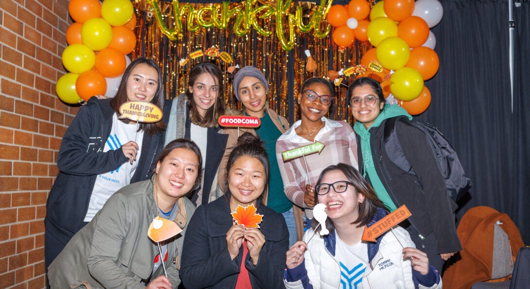 Eight smiling students pose in front of a gold and orange backdrop of streamers and balloons. A balloon displaying the word 