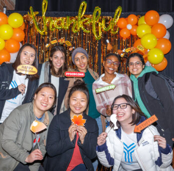Eight smiling students pose in front of a gold and orange backdrop of streamers and balloons. A balloon displaying the word 