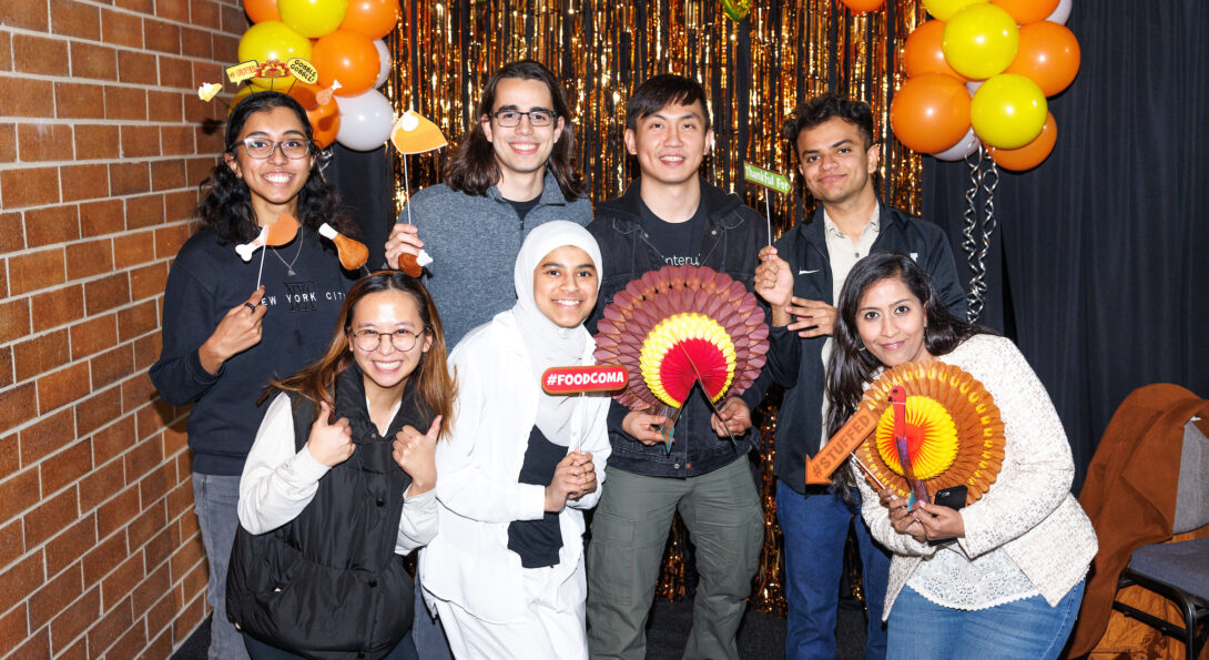 Eight students stand in front of a gold and yellow backdrop holding paper turkeys and other Thanksgiving-themed accessories.