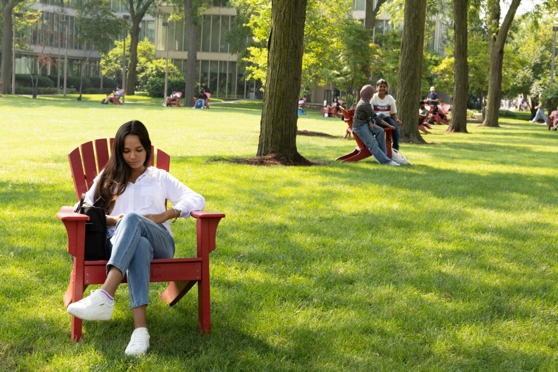 A student sits in a red aderondak chair in a grassy field on the UIC campus in summer.