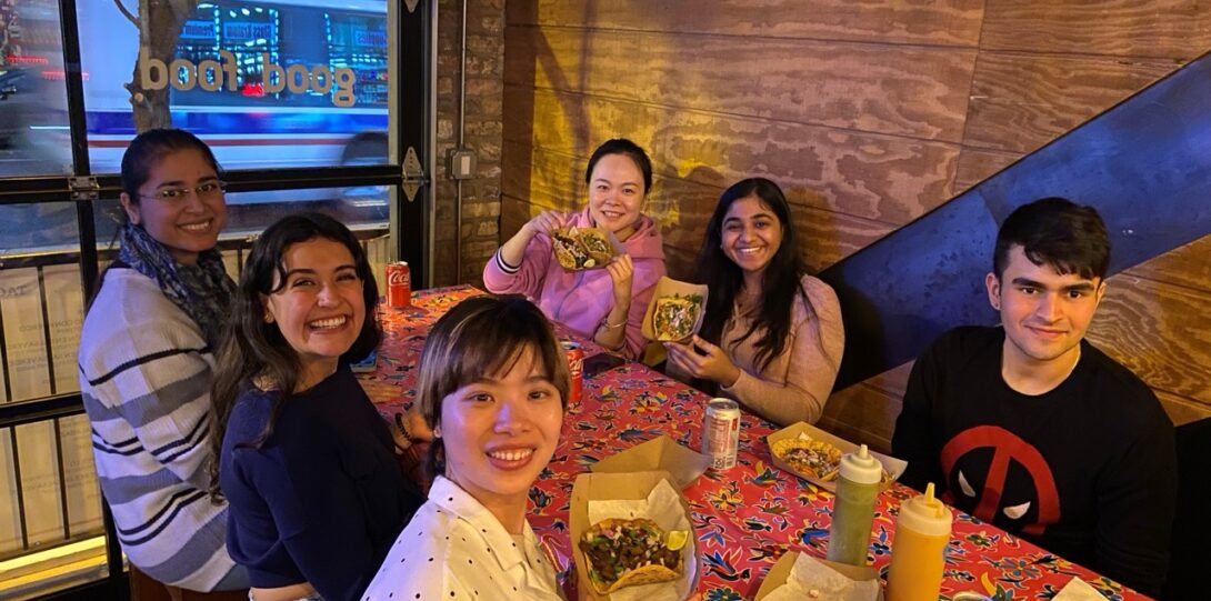 A group of students sit around a table holding up plates of tacos.