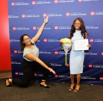 A student in a blue dress holding a certificate and flowers stands in front of a backdrop with UIC logos on it. Another person kneals next to her with arms outstretched. 