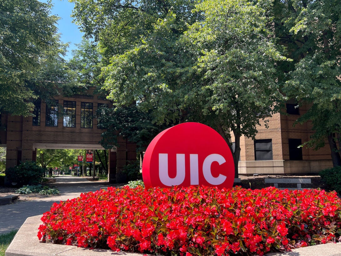 The UIC circle mark sculpture is shown on a sunny day with red flowers surrounding it. Green trees in the background.